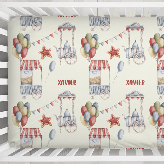 A crib mattress cover featuring a repeating watercolor pattern of carnival-themed illustrations, including balloons, popcorn stands, stars, bunting, and the personalized name 'XAVIER' in red. The cover is shown fitted inside a white wooden crib.