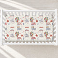 A top-down view of a white wooden crib containing a fitted sheet with a personalized circus-themed print. The sheet features illustrations of circus tents, balloons, bunting, and the name 'XAVIER' printed in red.