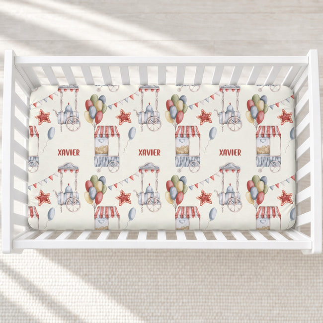 A top-down view of a white wooden crib containing a fitted sheet with a personalized circus-themed print. The sheet features illustrations of circus tents, balloons, bunting, and the name 'XAVIER' printed in red.