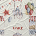 Personalized baby bedding or fabric featuring a watercolor fairground pattern with red, blue, and green balloons, a carousel, popcorn stand, and bunting on a cream background, with the name 'XAVIER' printed in red.