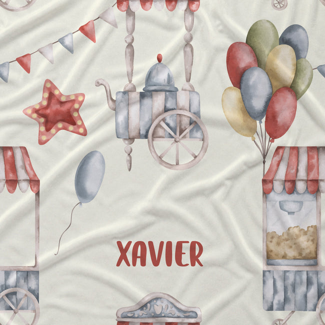 Personalized baby bedding or fabric featuring a watercolor fairground pattern with red, blue, and green balloons, a carousel, popcorn stand, and bunting on a cream background, with the name 'XAVIER' printed in red.