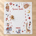 A personalized baby milestone blanket laid flat on a light wood floor. The white square blanket features circus-themed illustrations including a bear, lion, elephant, giraffe, and watercolor animals, with the name 'Leena Jade' and numbers 1 through 12 arranged in a border around 'MONTHS'.