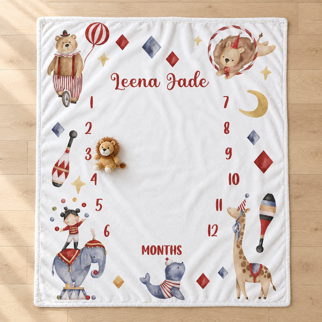 A personalized baby milestone blanket laid flat on a light wood floor. The white square blanket features circus-themed illustrations including a bear, lion, elephant, giraffe, and watercolor animals, with the name 'Leena Jade' and numbers 1 through 12 arranged in a border around 'MONTHS'.