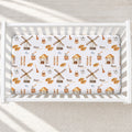 A white crib fitted with a printed baby bedding sheet featuring windmills, houses, ladders, and the name 'Knox William' repeated in a rustic pattern, photographed from above on a light textured floor.