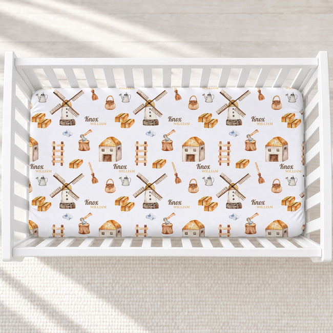 A white crib fitted with a printed baby bedding sheet featuring windmills, houses, ladders, and the name 'Knox William' repeated in a rustic pattern, photographed from above on a light textured floor.