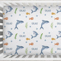 A custom baby crib mattress fitted sheet with an underwater theme, featuring gray and orange fish, green seaweed, blue bubbles, and the name 'SILAS' repeated on a white background.