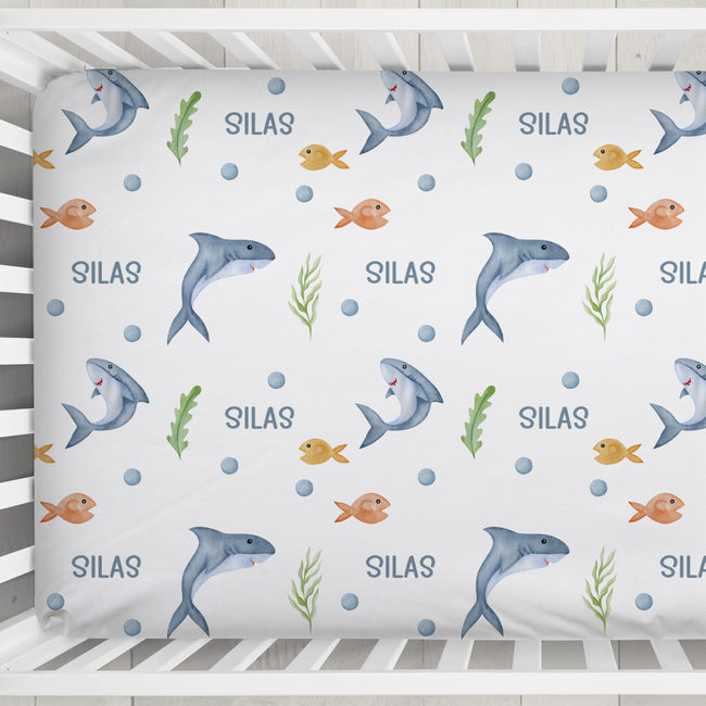 A custom baby crib mattress fitted sheet with an underwater theme, featuring gray and orange fish, green seaweed, blue bubbles, and the name 'SILAS' repeated on a white background.