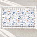 A white crib mattress fitted sheet with an underwater theme print featuring sharks, fish, bubbles, and the name 'SILAS' repeated, shown inside a white wooden crib.
