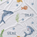 White fabric printed with a playful underwater pattern featuring blue-gray sharks, orange fish, green seaweed, blue circles, and the name 'SILAS' in blue text.