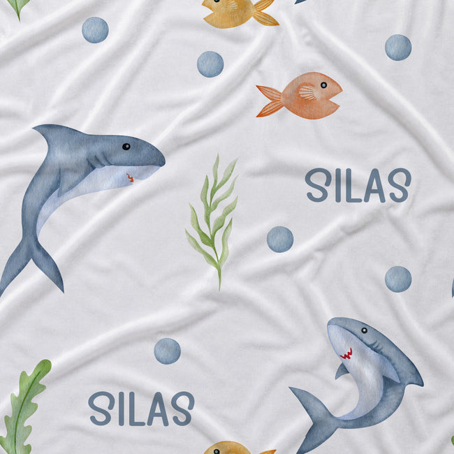 White fabric printed with a playful underwater pattern featuring blue-gray sharks, orange fish, green seaweed, blue circles, and the name 'SILAS' in blue text.