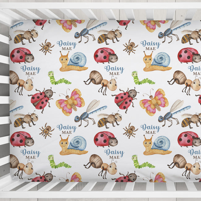 A crib bedding set featuring a white fitted sheet printed with a colorful pattern of cartoon ladybugs, butterflies, ants, caterpillars, and a snail, each accompanied by the text 'Daisy MAE'.