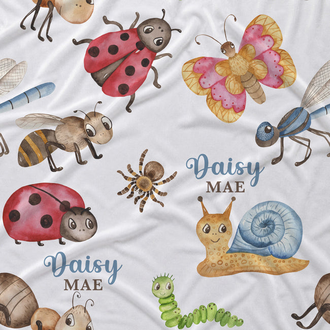 A close-up of a white fabric printed with a whimsical insect and nature pattern, including ladybugs, a butterfly, bees, a spider, a snail, a green caterpillar, and the personalized text 'Daisy MAE' in blue script.