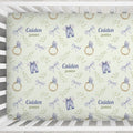 Custom baby crib sheet featuring a repeating pattern of watercolor blue baby outfits, gold rings, and green leaves on a pale yellow background, with personalized text 'Caiden James' printed in the center.