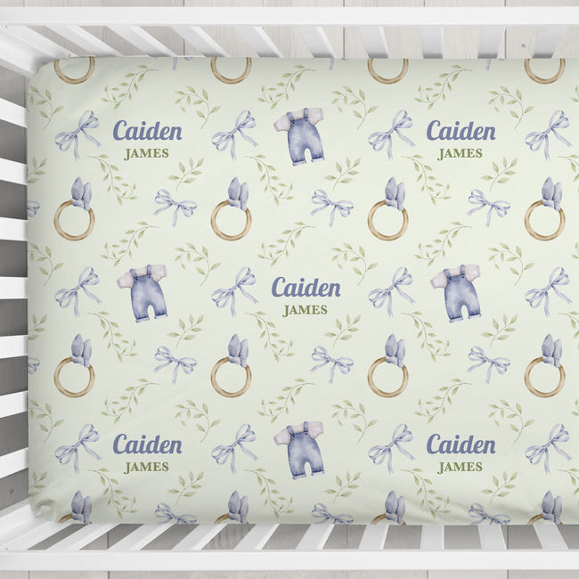 Custom baby crib sheet featuring a repeating pattern of watercolor blue baby outfits, gold rings, and green leaves on a pale yellow background, with personalized text 'Caiden James' printed in the center.