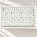 A personalized baby crib mattress cover with a repeating pattern of blue rabbits, gold rings, and green leaves on a cream background, shown inside a white wooden crib.