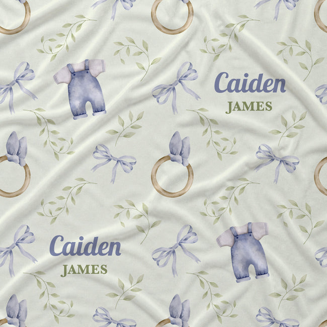 Personalized baby blanket featuring a repeating pattern of lavender baby overalls, wooden ring toys, lavender bows, and green leafy vines on a cream background, with the custom text 'Caiden JAMES' printed in blue and olive green.