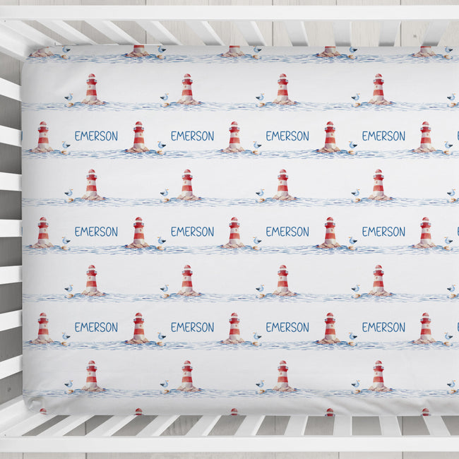 Personalized baby bedding set in a white crib, featuring a repeating pattern of red and white lighthouses, blue seagulls, and the name 'EMERSON' printed in blue.