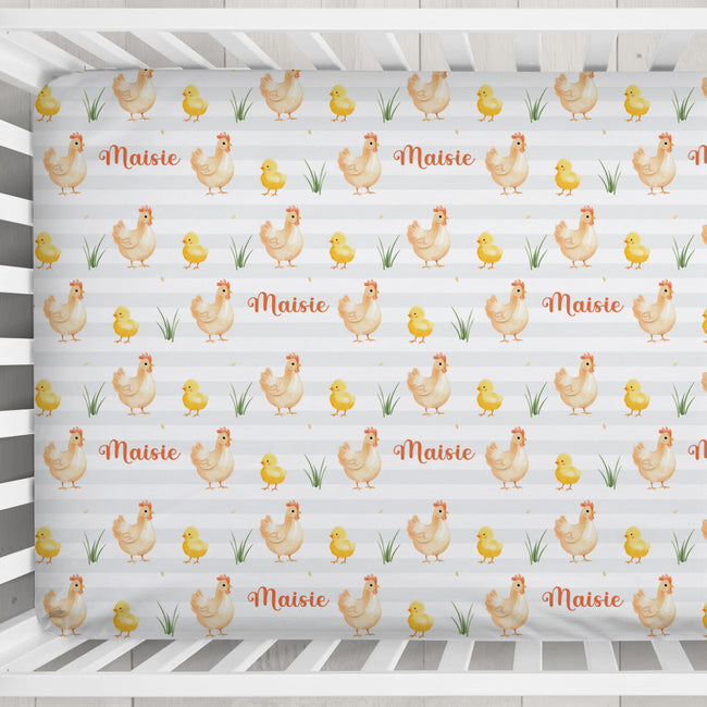 A fitted crib sheet with a repeating pattern of cartoon chickens, chicks, grass, and the name 'Maisie' in orange script, shown inside a white wooden crib.