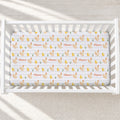 A white crib mattress fitted with a repeating chicken and chick print sheet, shown inside a white wooden crib on a light neutral floor with soft window shadows.