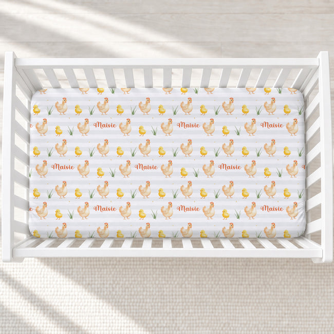 A white crib mattress fitted with a repeating chicken and chick print sheet, shown inside a white wooden crib on a light neutral floor with soft window shadows.