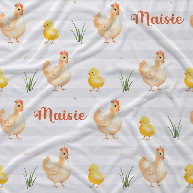 A close-up of a fabric surface with a repeating Easter-themed print featuring watercolor-style chickens, yellow chicks, green grass, and the name 'Maisie' in orange script on a white background.