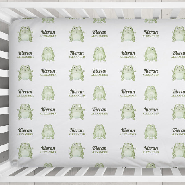 A crib mattress fitted sheet with a repeating pattern of light green cartoon frogs and the custom text 'Kieran ALEXANDER' on a white background, shown inside a white crib.