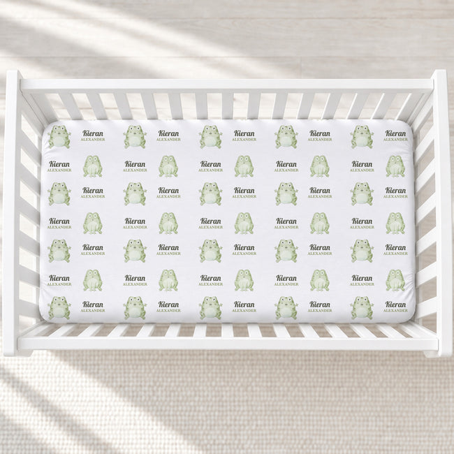 A white crib mattress fitted sheet with a repeating pattern of light green cartoon frogs and the name 'Kieran Alexander' printed in dark gray, shown inside a white wooden crib.