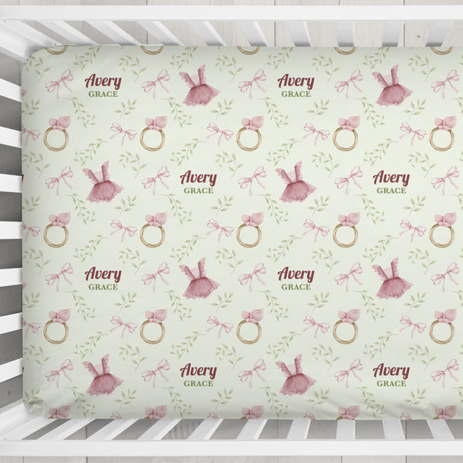 A personalized baby crib sheet featuring a repeating pattern of pink tutus, pink bows, gold rings, and green leaves on a pale sage green background. The text 'Avery GRACE' is printed in a dusty rose font, indicating a custom name combination.