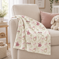 A cream-colored baby blanket with pink bows, green leaves, and gold rings, draped over the arm of a beige armchair in a nursery setting.