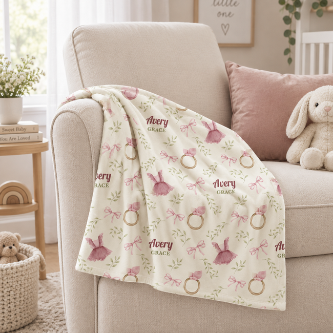 A cream-colored baby blanket with pink bows, green leaves, and gold rings, draped over the arm of a beige armchair in a nursery setting.