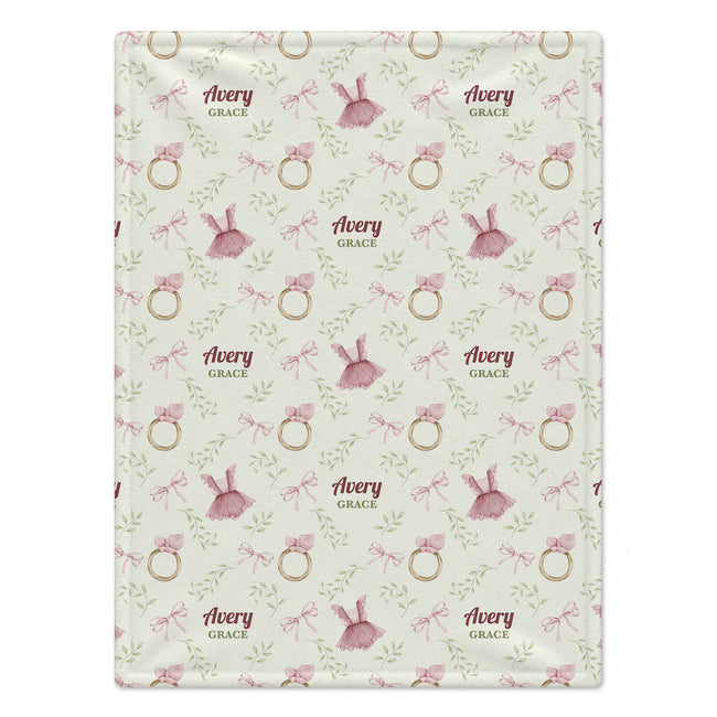 A personalized baby blanket with a repeating pattern of pink bows, gold rings, green leaves, and pink dresses, with the custom text 'Avery Grace' printed in dark red. The blanket is laid flat against a clean white background.
