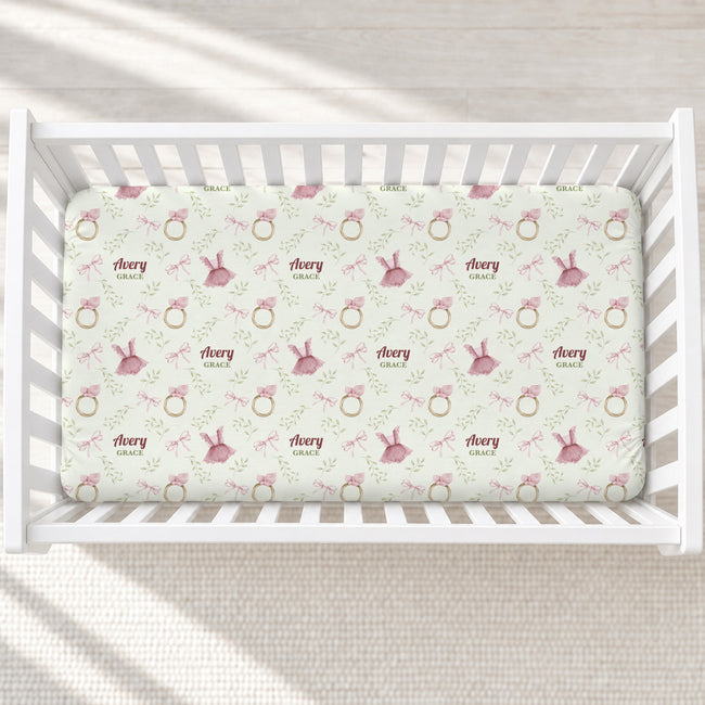 A top-down view of a white wooden crib containing a fitted crib mattress with a repeating pattern of pink bunnies, gold rings, and the text 'Theory Grace' on a light cream background.