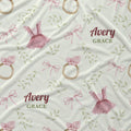 A fabric or wallpaper pattern featuring pink ballerina dresses, gold rings, pink bows, and green leaves on a cream background, with the personalized text 'Avery GRACE' printed in maroon and olive.