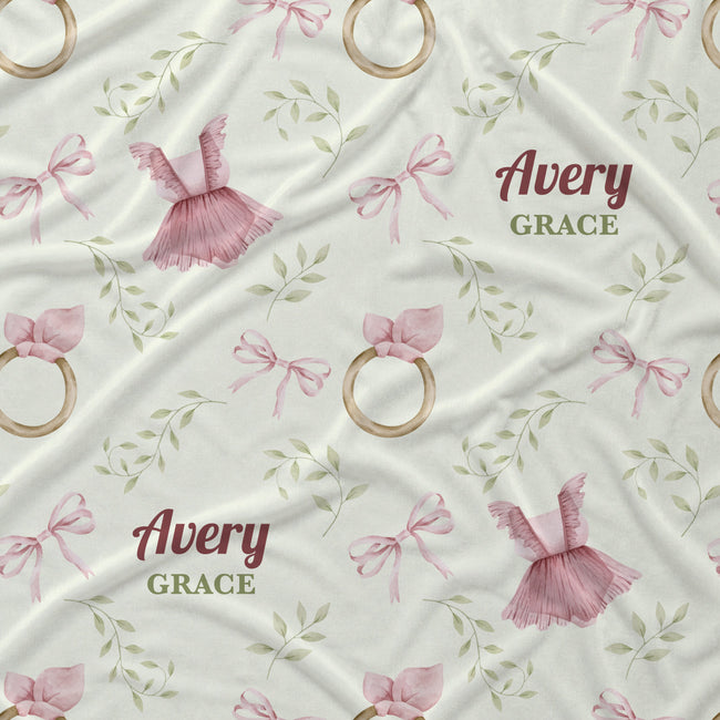 A fabric or wallpaper pattern featuring pink ballerina dresses, gold rings, pink bows, and green leaves on a cream background, with the personalized text 'Avery GRACE' printed in maroon and olive.