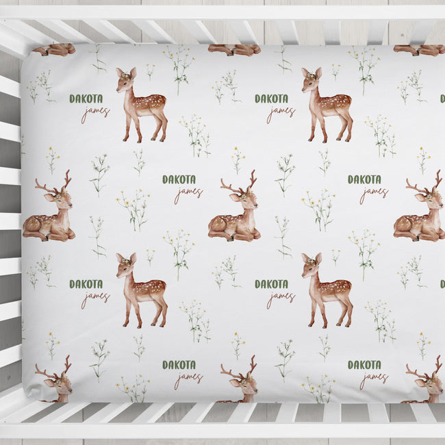 A personalized crib sheet with a repeating woodland print featuring brown deer illustrations, small wildflower motifs, and the words 'Dakota James' in brown script.