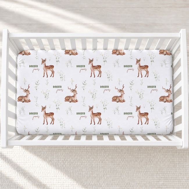 A white crib mattress fitted with a printed sheet featuring brown deer illustrations, small green leaves, and the names 'Dakota James' repeated across the surface, shown in a white crib on a light textured floor.
