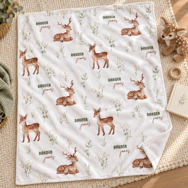 A white fleece blanket with an all-over print of brown deer, small white flowers, and the words 'Dakota james' in green and brown, styled on a woven beige rug with a plush toy and small props nearby.