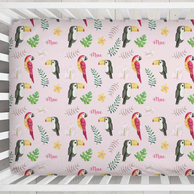 A crib sheet covered in a tropical pattern featuring toucans, green leaves, small yellow flowers, and the name 'Mae' in pink script, shown inside a white wooden crib.