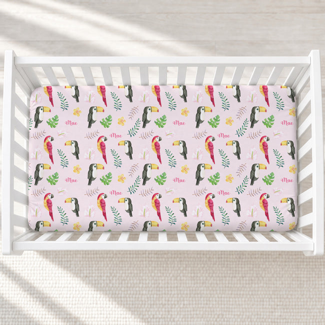 A fitted crib sheet with a colorful toucan and tropical leaf pattern on a light pink background, shown inside a white wooden crib.