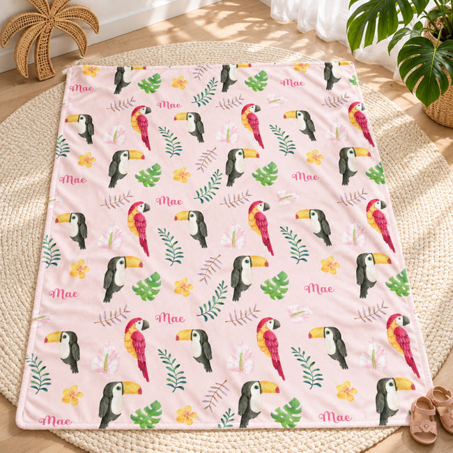 Personalized Tropical Bird Baby Girl Blanket. Colorful parrots, toucans, tropical leaves, and flowers surround a custom name on a soft pink background.