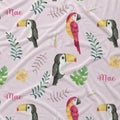 A close-up of a light pink fabric with a tropical print featuring watercolor-style toucans, a red and yellow parrot, green leaves, pink flowers, and the name 'Mae' repeated in pink script.
