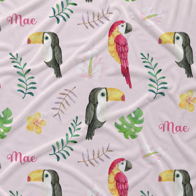 A close-up of a light pink fabric with a tropical print featuring watercolor-style toucans, a red and yellow parrot, green leaves, pink flowers, and the name 'Mae' repeated in pink script.