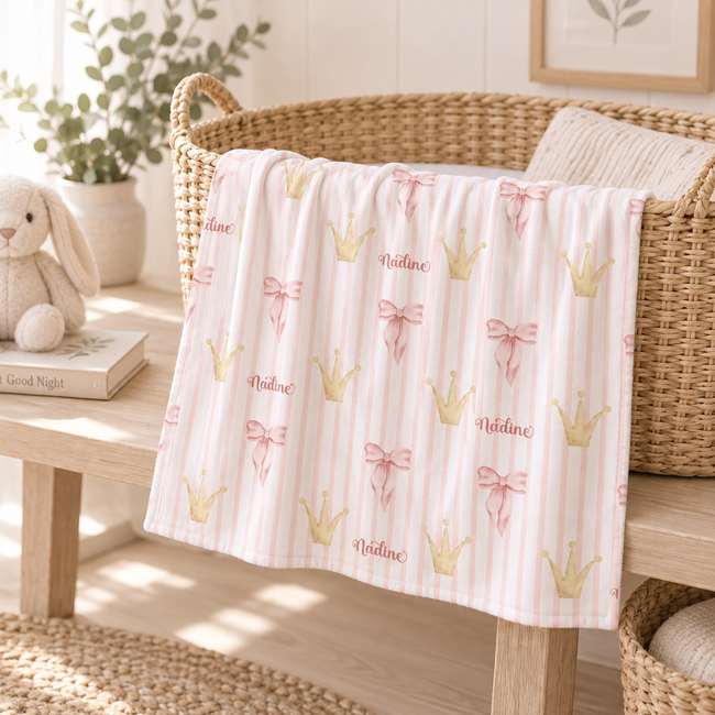 A baby blanket draped over a wicker basket in a nursery setting. The blanket has a light pink and white vertical stripe pattern with gold crowns, pink bows, and the name 'Nadine' printed in script.