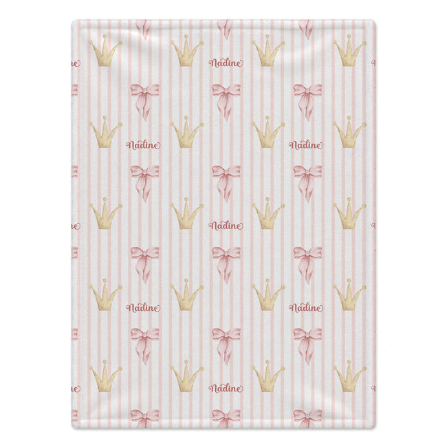A rectangular fabric item, likely a blanket or towel, featuring a repeating pattern of gold crowns, pink bows, and the name 'Nadine' on a light pink and white striped background.