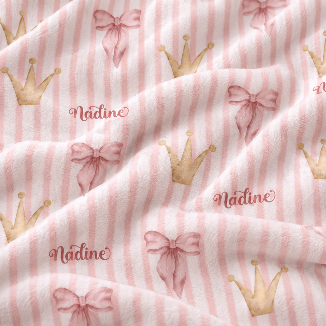 A close-up of a soft plush fabric with a pattern of light pink and white vertical stripes, decorated with watercolor-style pink bows, gold crowns, and the name 'Nadine' in cursive.
