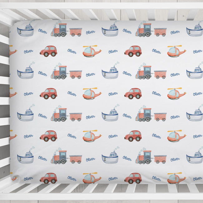 A baby crib mattress fitted sheet with a repeating pattern of colorful toy vehicles (trains, boats, cars, helicopters) and the name 'Oliver' printed in blue script, shown inside a white wooden crib.