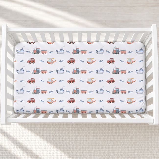 A fitted crib mattress with a white background and a repeating pattern of colorful toy vehicles, including cars, boats, helicopters, and tractors, shown inside a white wooden crib.