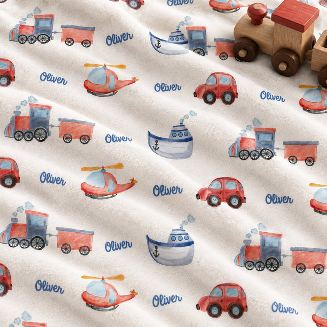 Personalized baby fabric with a playful watercolor pattern of trains, helicopters, cars, boats, and the name 'Oliver' printed in blue script, shown with a wooden toy train on top.