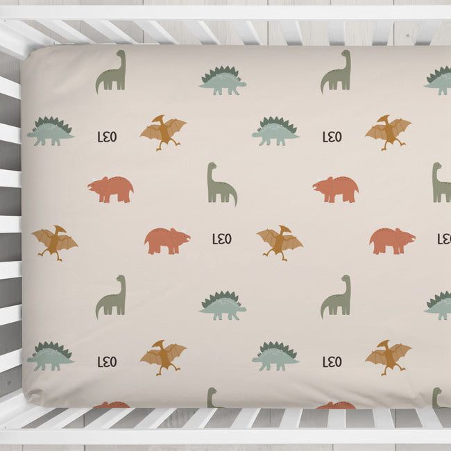 A fitted crib sheet with a dinosaur pattern, shown inside a white wooden crib. The sheet features a repeating pattern of green, orange, and brown dinosaurs on a warm beige background, with the name 'LEO' printed in black between some motifs.
