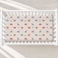 A fitted crib sheet with a dinosaur pattern in muted green, orange, and brown on a light blush background, shown inside a white wooden crib.